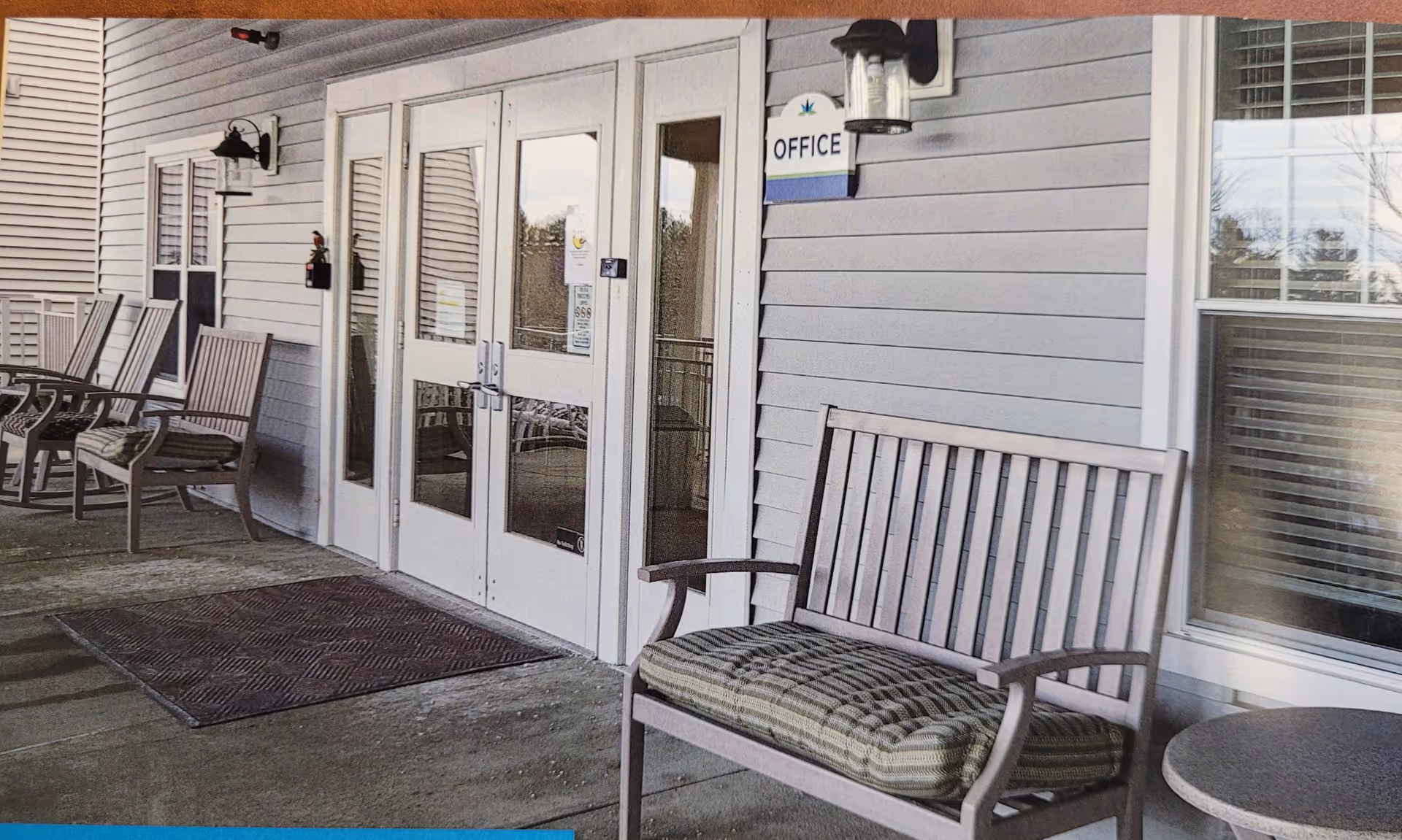 Outdoor covered patio area with wooden chairs and benches with cushions, a small round table, and double glass doors labeled 'Office'. The building exterior is light gray with white trim and has wall-mounted lantern-style lights.