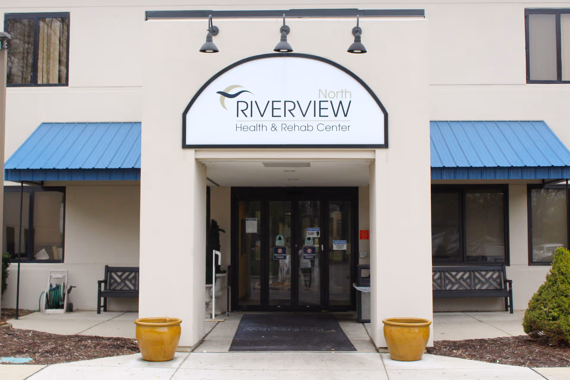 Front entrance of Riverview North Health & Rehab Center showing the building sign above double glass doors, blue awnings, benches and two yellow planters.
