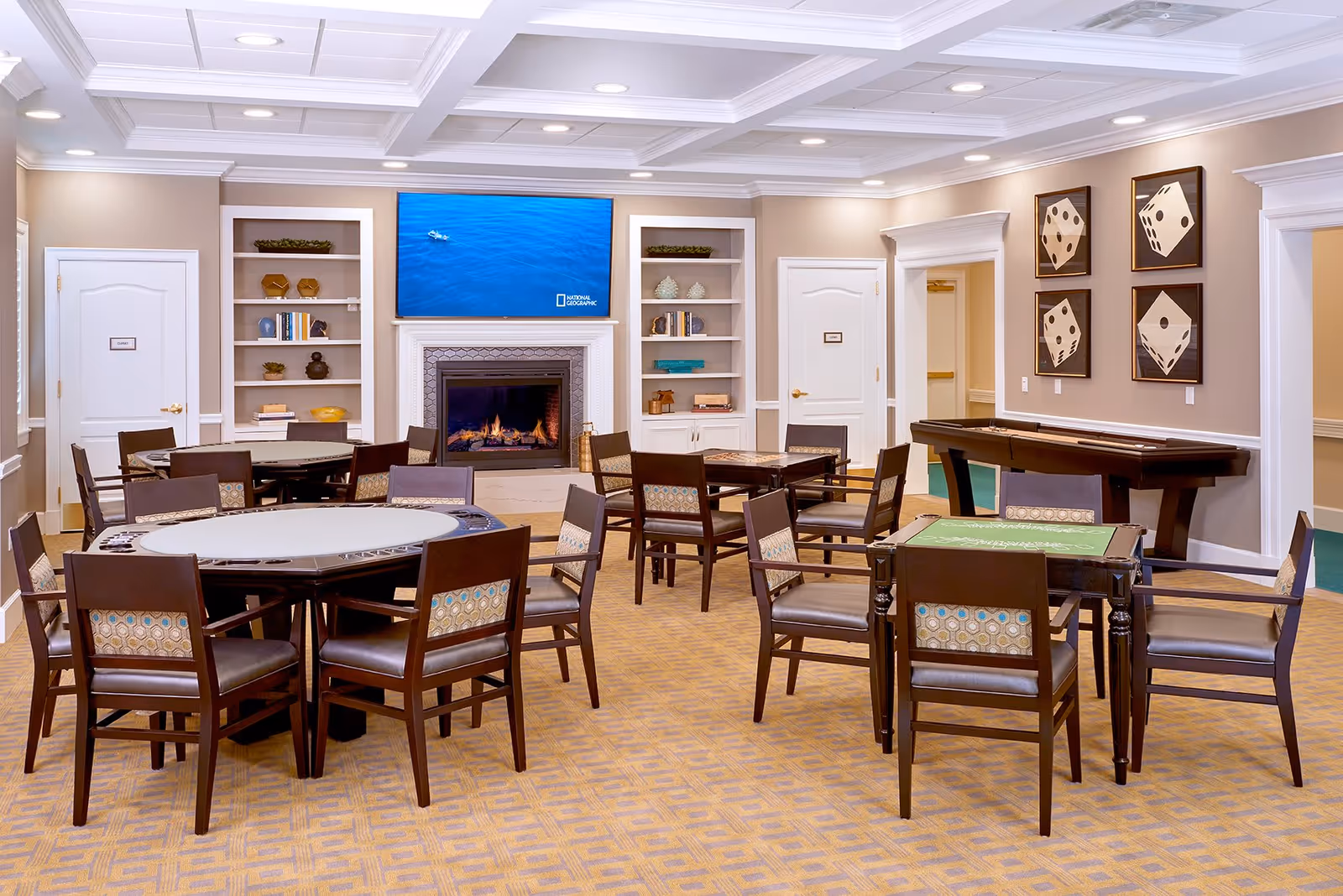 A well-lit common room with multiple tables and chairs arranged for group activities. The room features a fireplace with a flat-screen TV above it displaying a National Geographic program. Built-in shelves with decorative items flank the fireplace. On the right wall, there are four framed pictures of dice. The room has a patterned carpet and a shuffleboard table along the right wall.