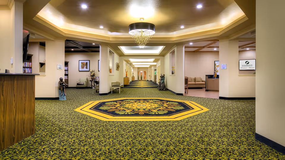 A spacious, well-lit hallway in an assisted living facility with patterned green and yellow carpet, decorative statues, and seating areas visible on the sides. The ceiling features recessed lighting and a central chandelier.