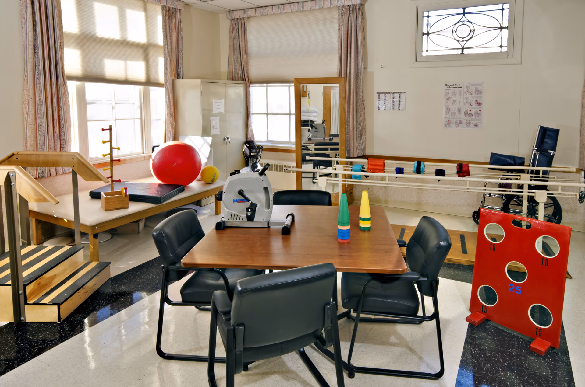 A rehabilitation therapy room with exercise equipment including a stationary bike, therapy stairs, balance bars, a large red exercise ball, and a table with four black chairs. The room has large windows with curtains and a mirror on the wall. There is also a red target board with holes and a wheelchair in the background.