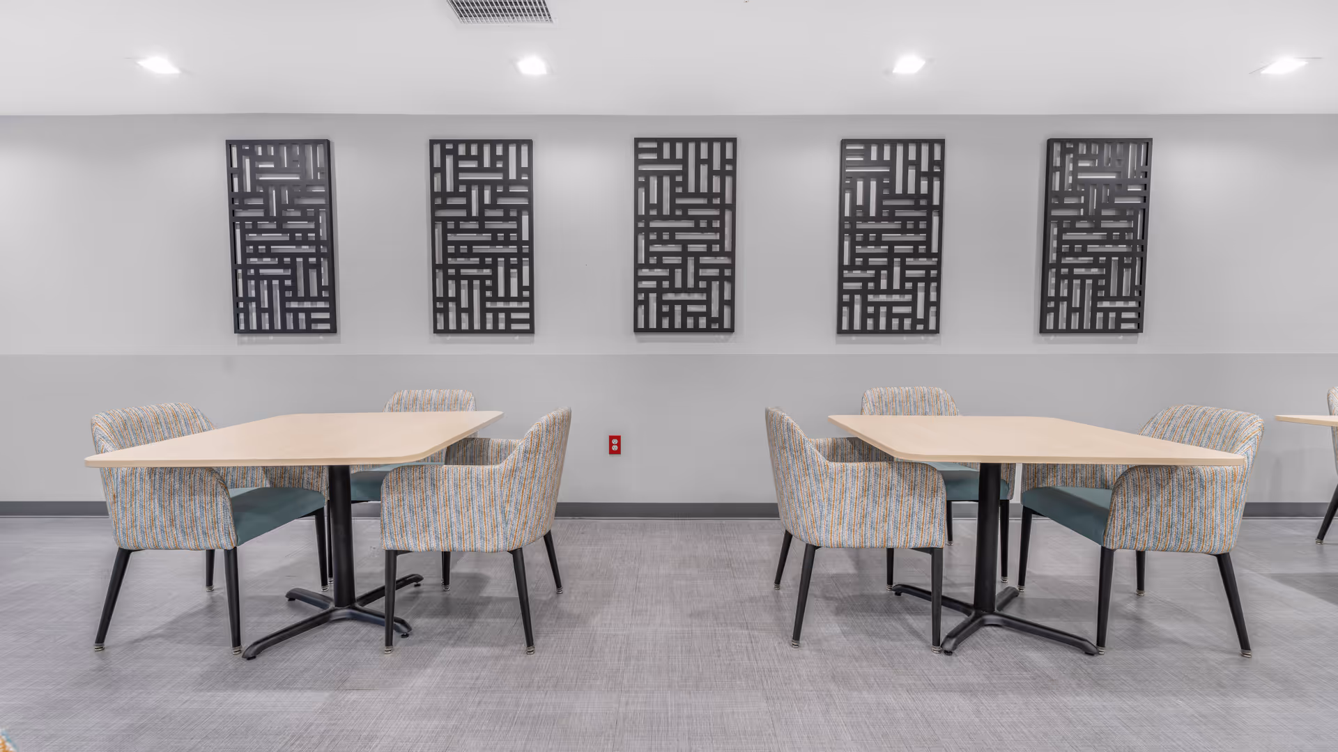 A clean and modern dining area with two light wood rectangular tables, each surrounded by four upholstered chairs with patterned fabric and black legs. The walls are painted white with a gray lower half, and five decorative black geometric wall panels are evenly spaced above the tables. The floor is covered with gray tiles, and ceiling lights provide bright illumination.