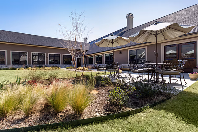 Outdoor patio area at Briarview Senior Living with metal tables and chairs under large umbrellas, surrounded by landscaped garden beds with ornamental grasses and shrubs, adjacent to a single-story building with multiple windows.