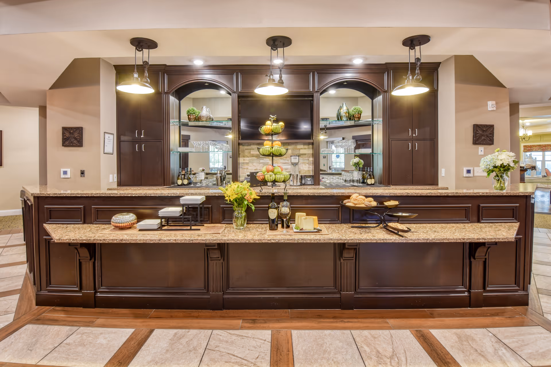 A well-lit kitchen or serving area with dark wood cabinetry and granite countertops. The counter displays a tiered fruit stand with apples and pears, bottles of wine, a cheese platter, flowers in a vase, and plates with pastries. There are three pendant lights hanging from the ceiling above the counter, and a large mirror with shelves holding glassware and decorative items in the background.