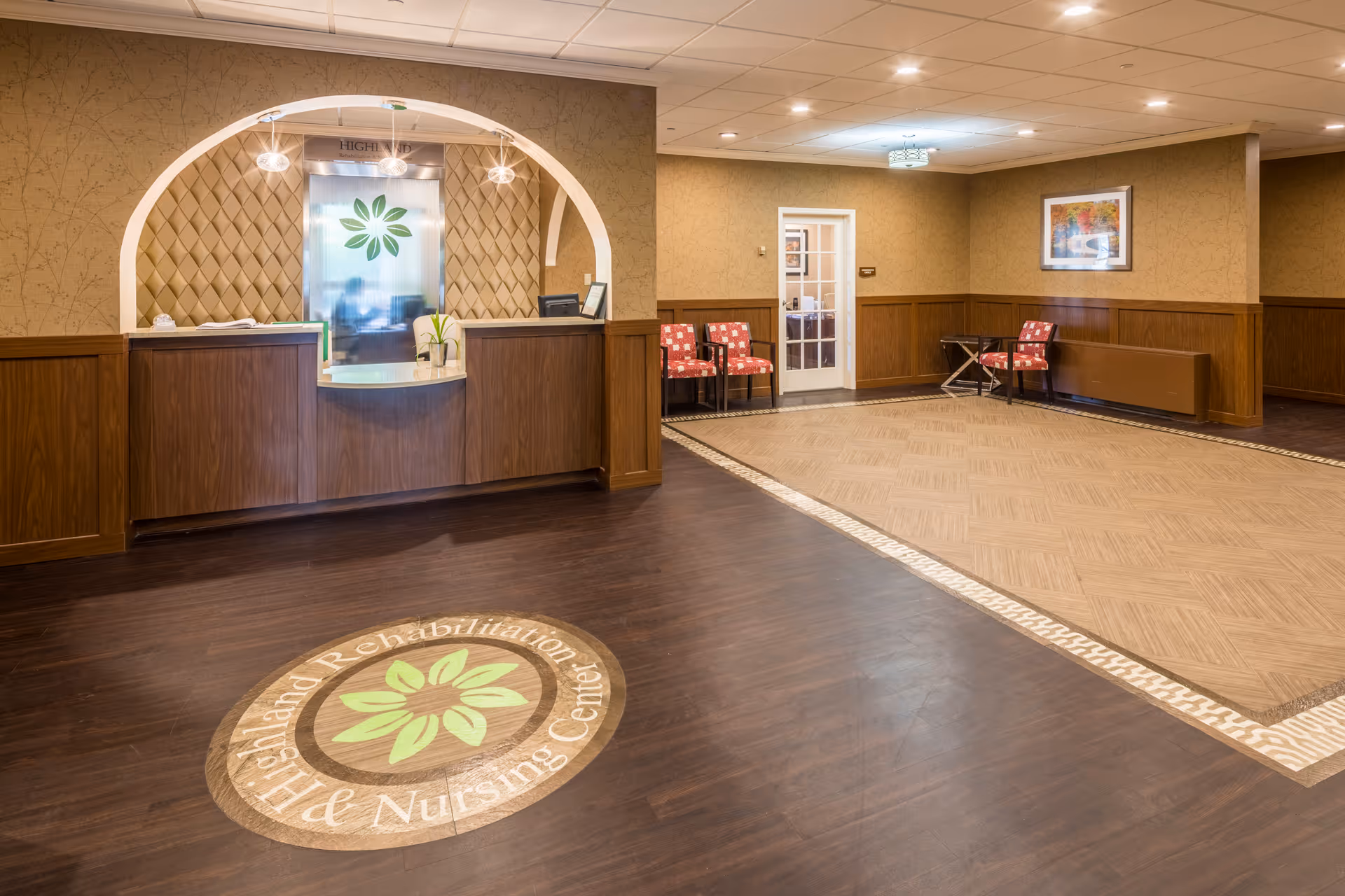 A spacious reception lobby with a wooden front desk, seating area, and a circular floor logo for Highland Rehabilitation & Nursing Center.