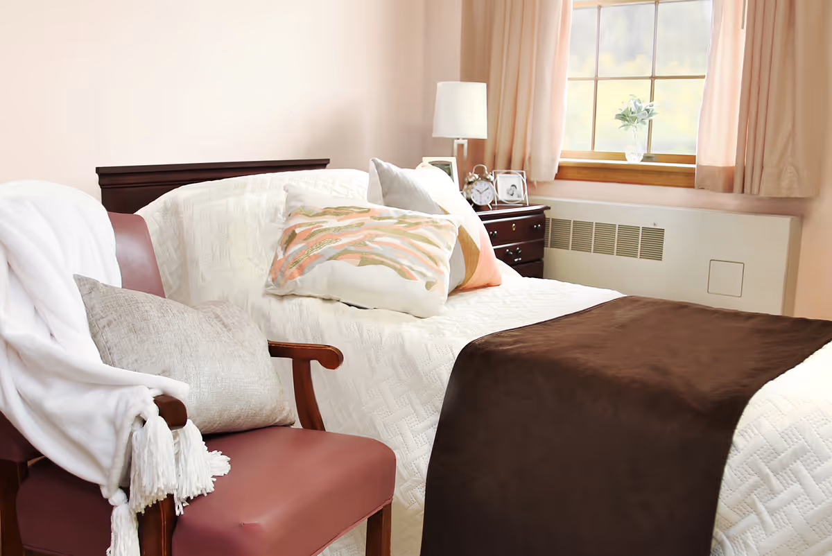 A neatly made single bed with decorative pillows and a brown throw next to a window and a cushioned chair draped with a blanket.