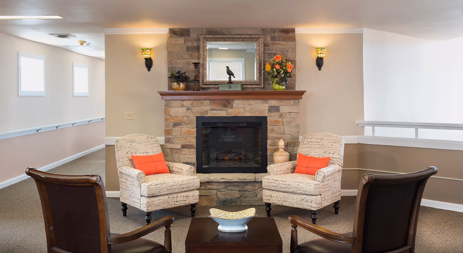 A cozy sitting area with a stone fireplace in the center, flanked by two beige armchairs with script patterns and orange cushions. Two dark wooden chairs face the armchairs across a small wooden coffee table with a decorative bowl. The walls are light-colored with two wall sconces on either side of the fireplace, and a large mirror and flower vase are on the mantel.