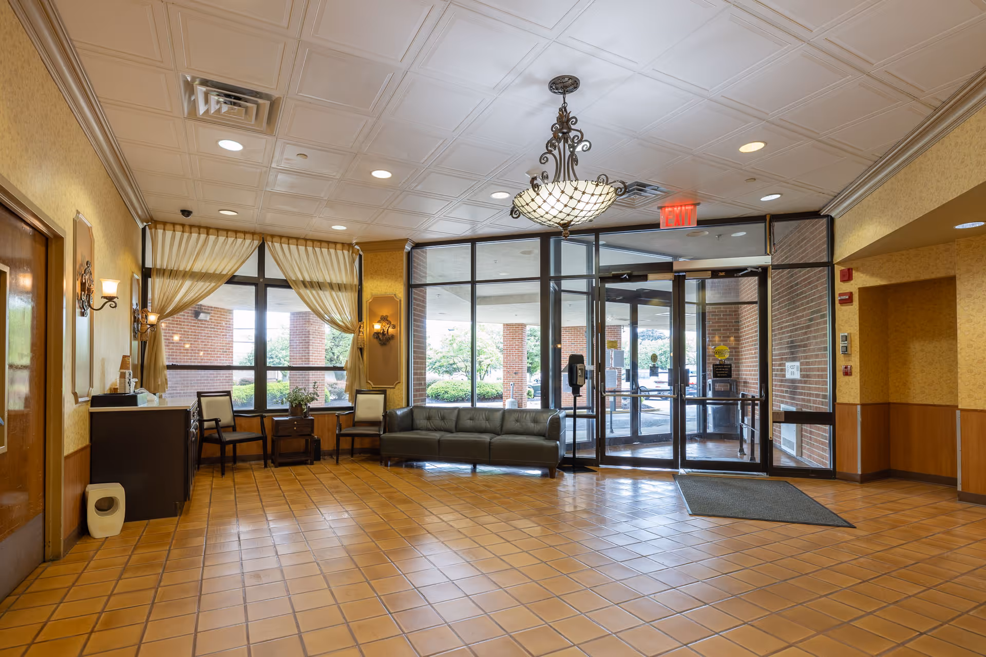 A spacious facility lobby with tiled floor, a chandelier hanging from the ceiling, a gray sofa, two chairs with a small table between them, large windows with curtains, and glass doors leading outside. The walls are decorated with sconces and light yellow wallpaper.