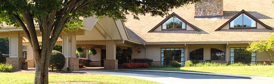Exterior view of Bermuda Village Retirement Center showing a single-story building with a covered entrance, large windows, and a sloped roof with dormer windows. There is a tree and well-maintained landscaping in the foreground.