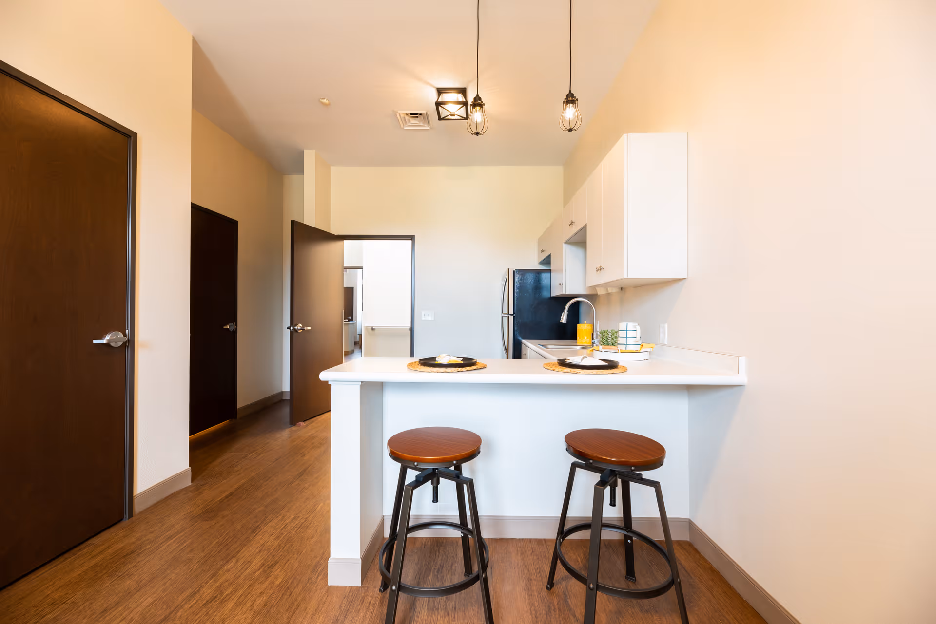 Interior view of a modern kitchen area in an assisted living facility with a white countertop bar, two brown stools, white cabinets, a black refrigerator, and wooden doors leading to other rooms.