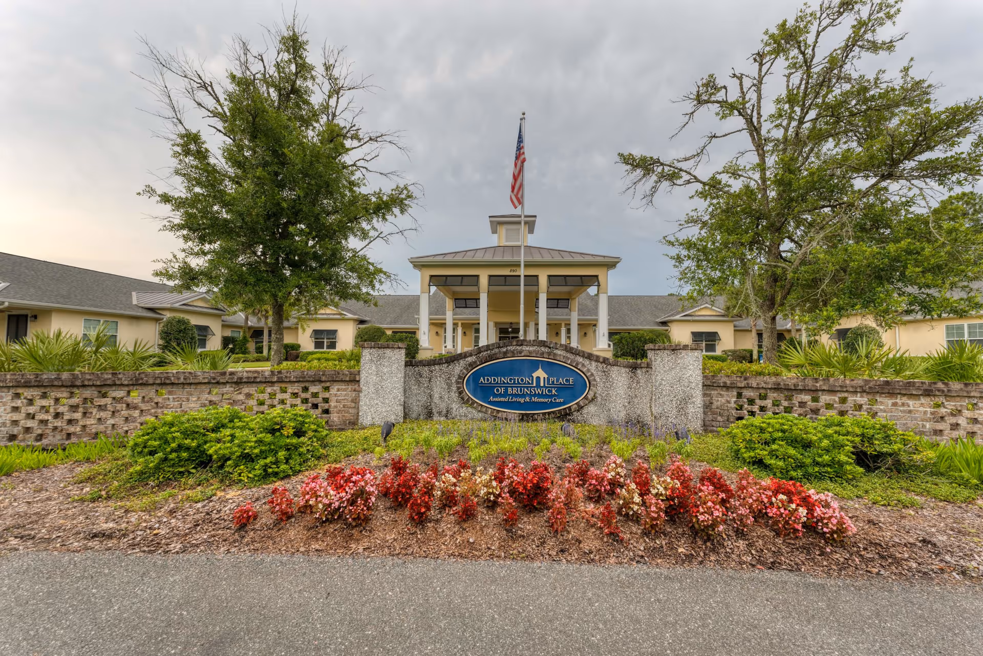 Front exterior view of Addington Place of Brunswick building with a landscaped garden featuring red and pink flowers, green shrubs, and two trees. A flagpole with an American flag stands in front of the building entrance.