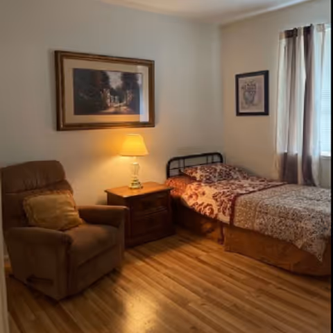 A small bedroom with a single bed, nightstand and lamp, a recliner chair, framed wall art, and hardwood floors.