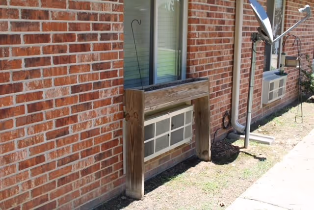 Exterior view of a brick building wall with two windows, each with an air conditioning unit installed below. There is a wooden frame structure around one of the air conditioning units and a satellite dish mounted on a pole near the building. The ground is bare soil with some grass patches and a concrete walkway beside the building.