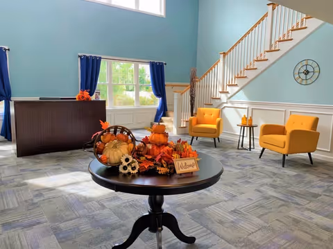 A bright and welcoming senior living facility lobby with light blue walls, a large window with blue curtains, two mustard yellow armchairs, a small side table with candles, and a round table in the center decorated with autumn-themed items including pumpkins, leaves, and a 'Welcome' sign. A staircase with wooden handrails is visible in the background.