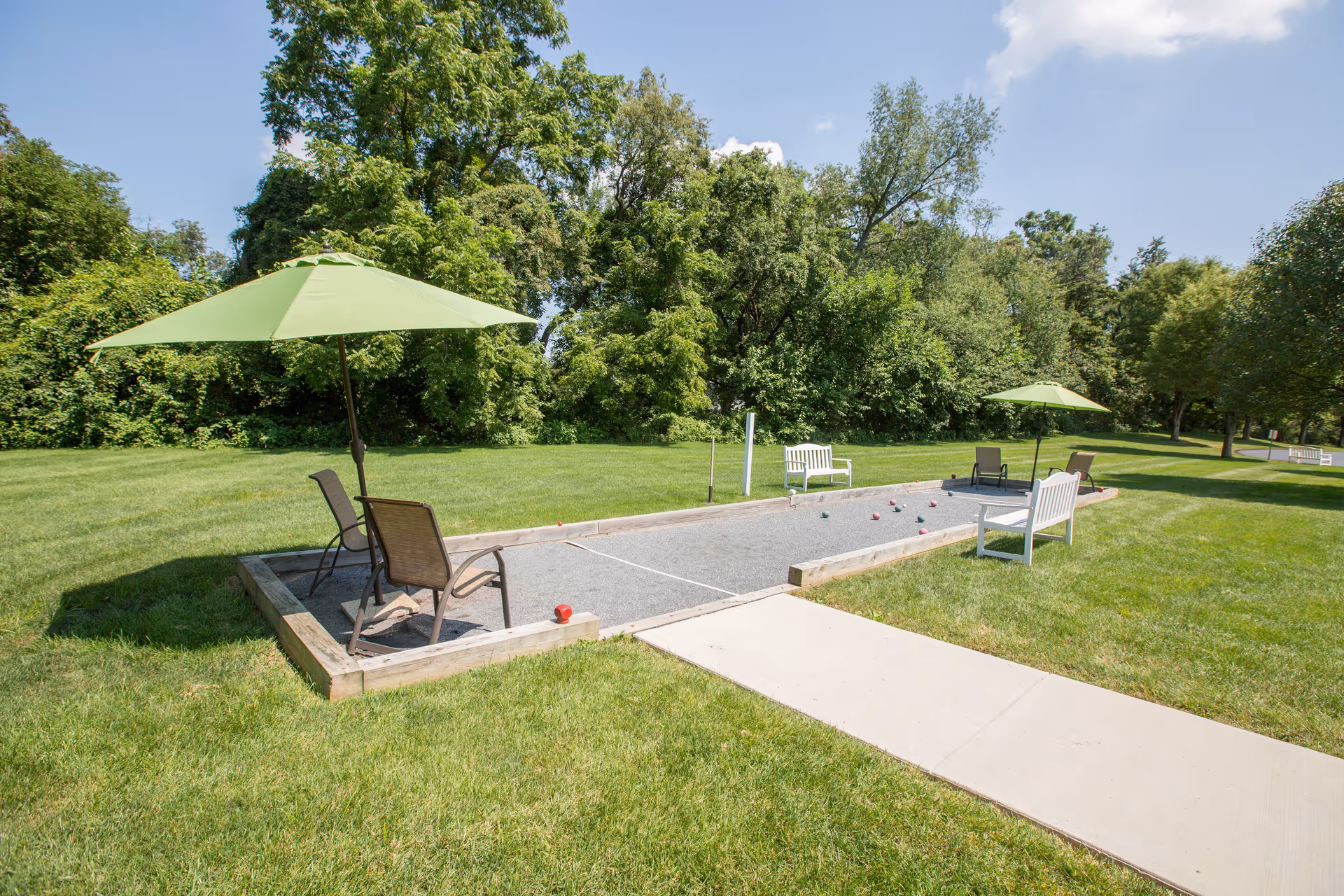 Outdoor bocce ball court with green umbrellas, chairs, and benches surrounded by green grass and trees under a blue sky with some clouds.
