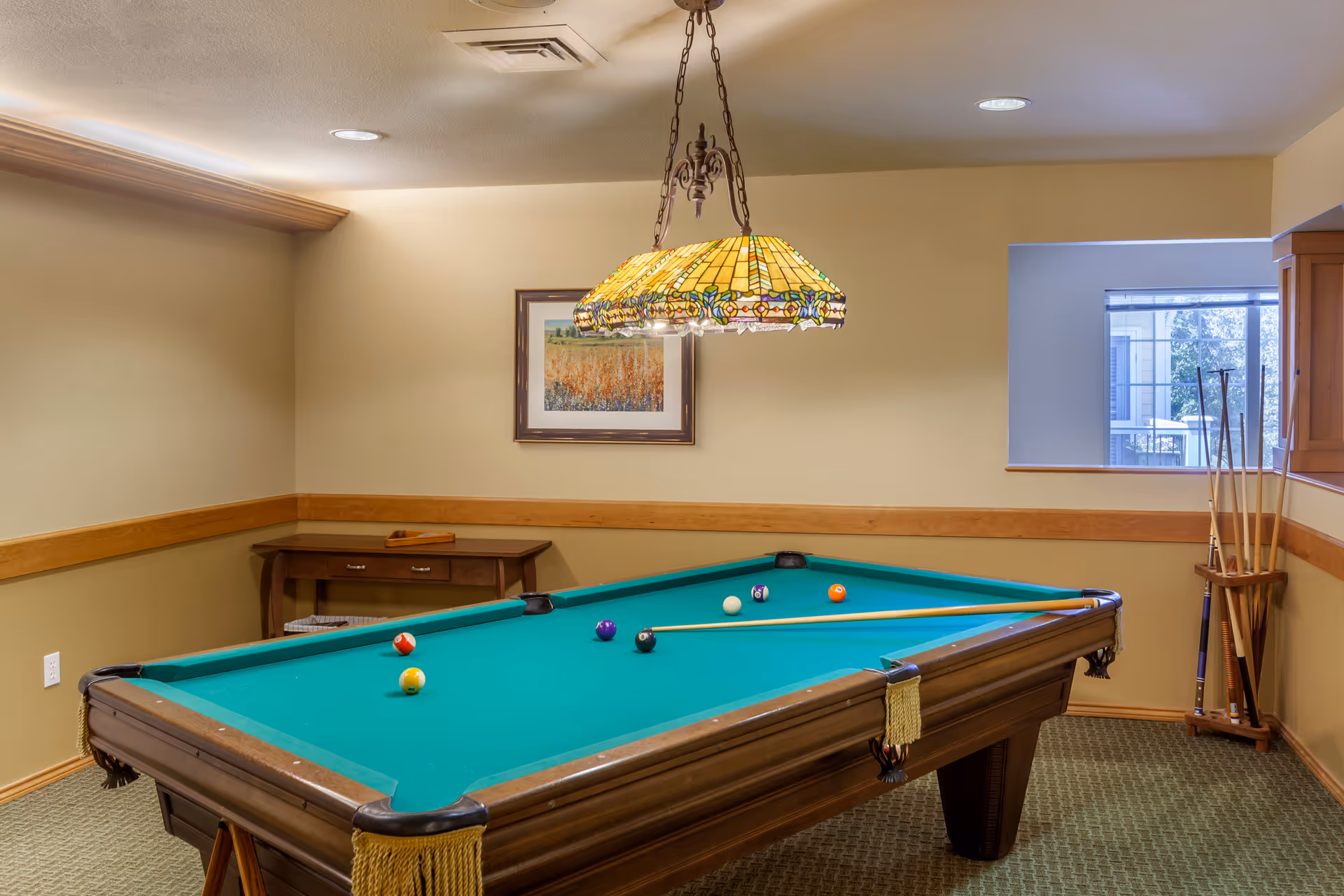 A billiards room with a pool table, hanging stained-glass lamp, cue rack, and framed artwork on the wall.