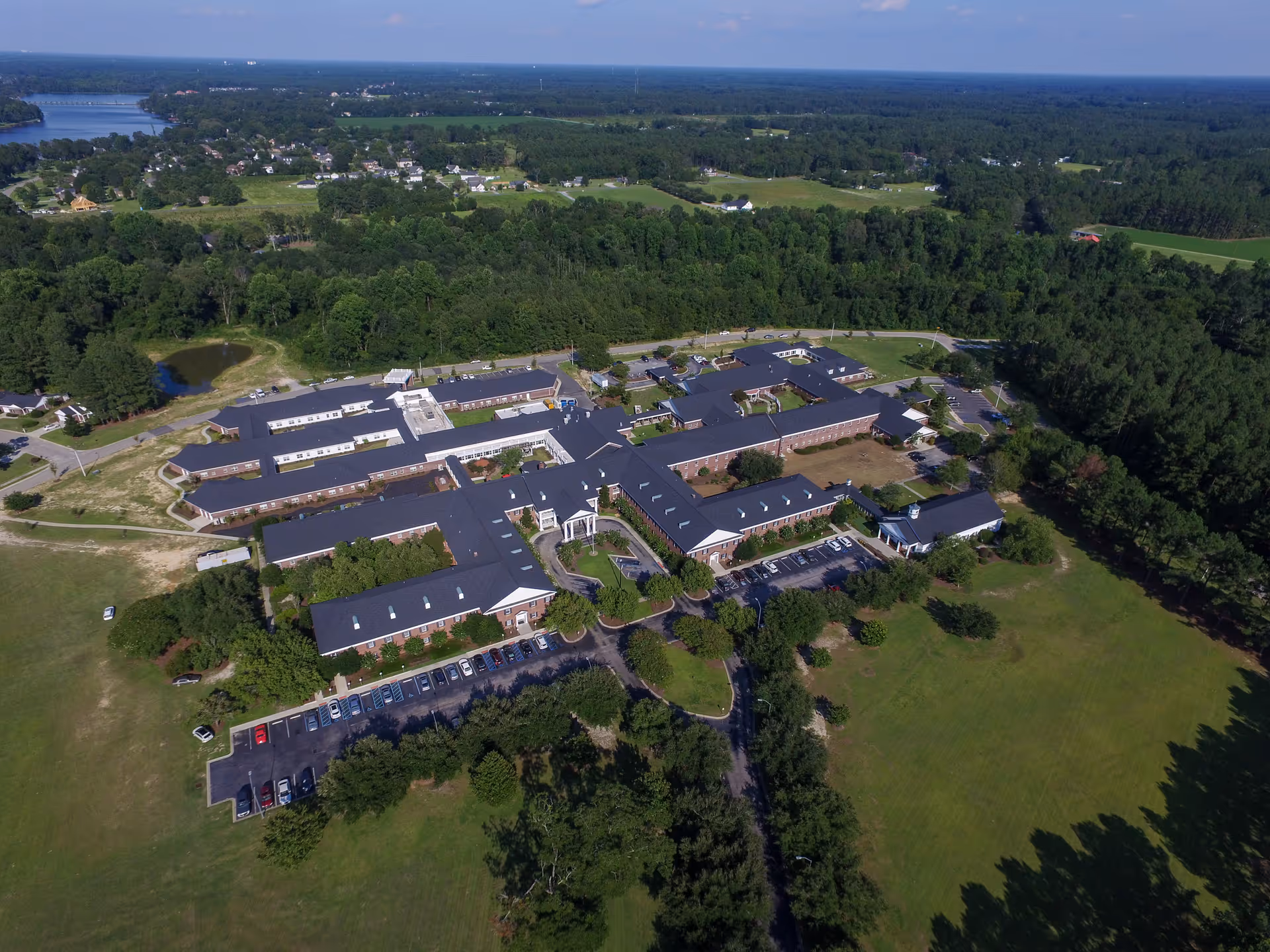 Aerial view of The Manor senior living facility surrounded by trees and greenery, with multiple connected buildings, parking lots, and open grassy areas.
