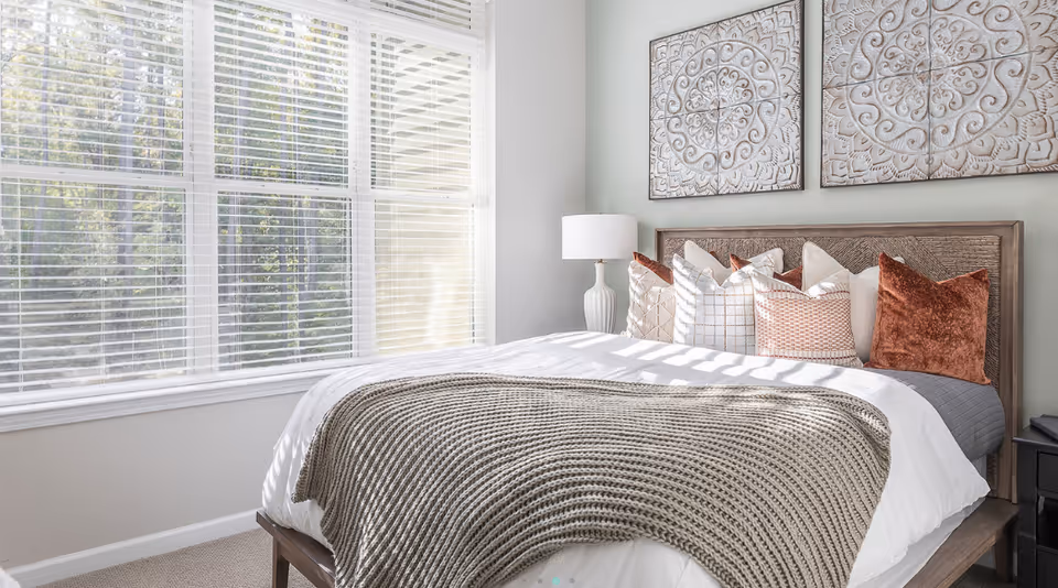 Sunlit bedroom with a made bed topped by a textured throw and decorative pillows, a bedside lamp, and a large window with blinds.