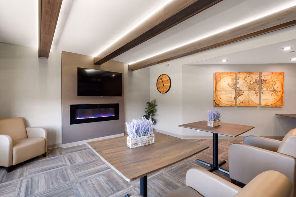 Bright communal living room with a wall-mounted TV above an electric fireplace, armchairs, tables with lavender centerpieces, a wall clock and world map artwork.