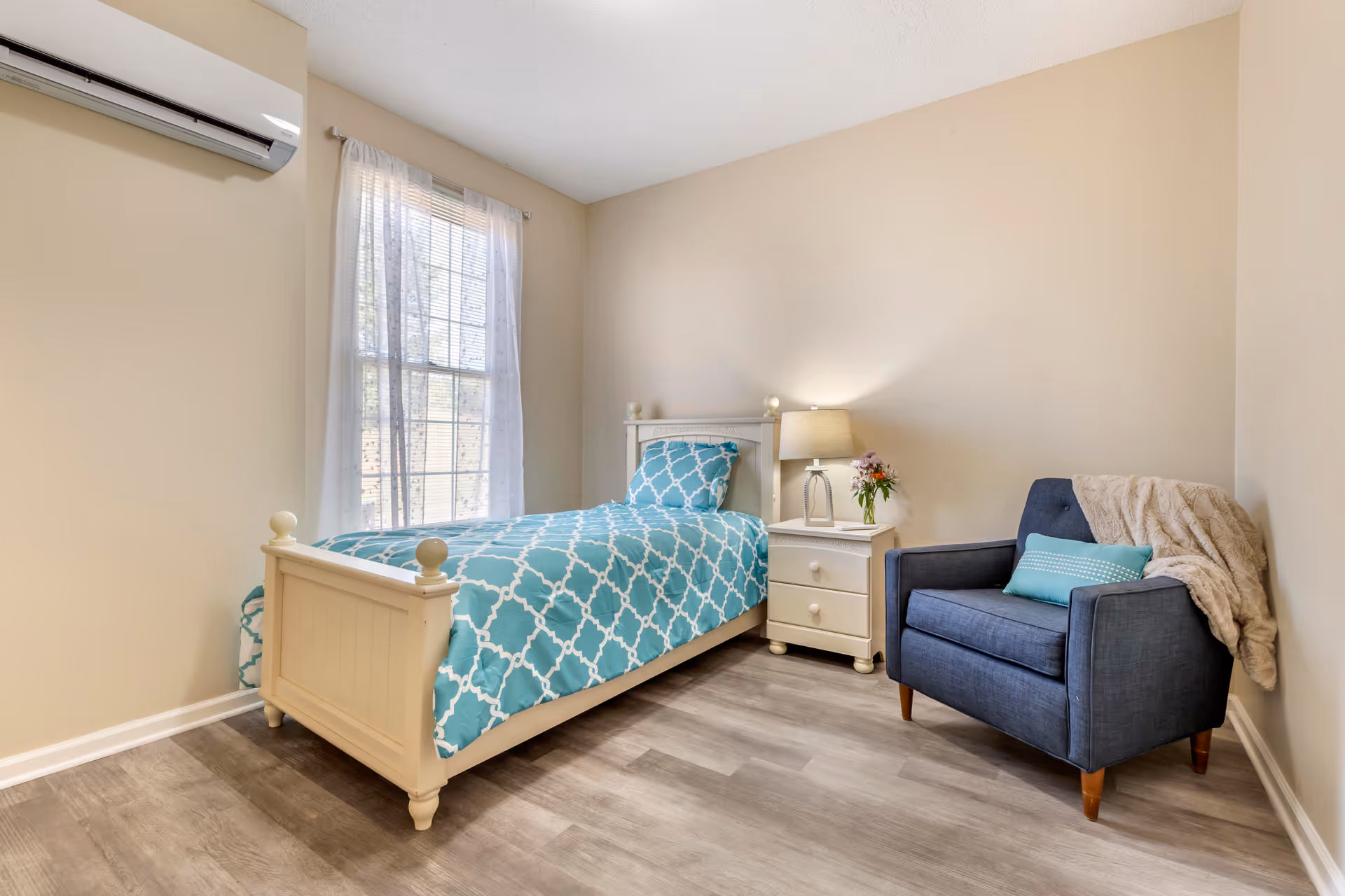 A small bedroom with a single bed featuring a blue and white patterned bedspread and pillow. Next to the bed is a white nightstand with a lamp and a small vase of flowers. A blue armchair with a teal pillow and a beige throw blanket is positioned in the corner. The room has light beige walls, a window with sheer white curtains, and wood-look flooring. An air conditioning unit is mounted on the wall above the window.