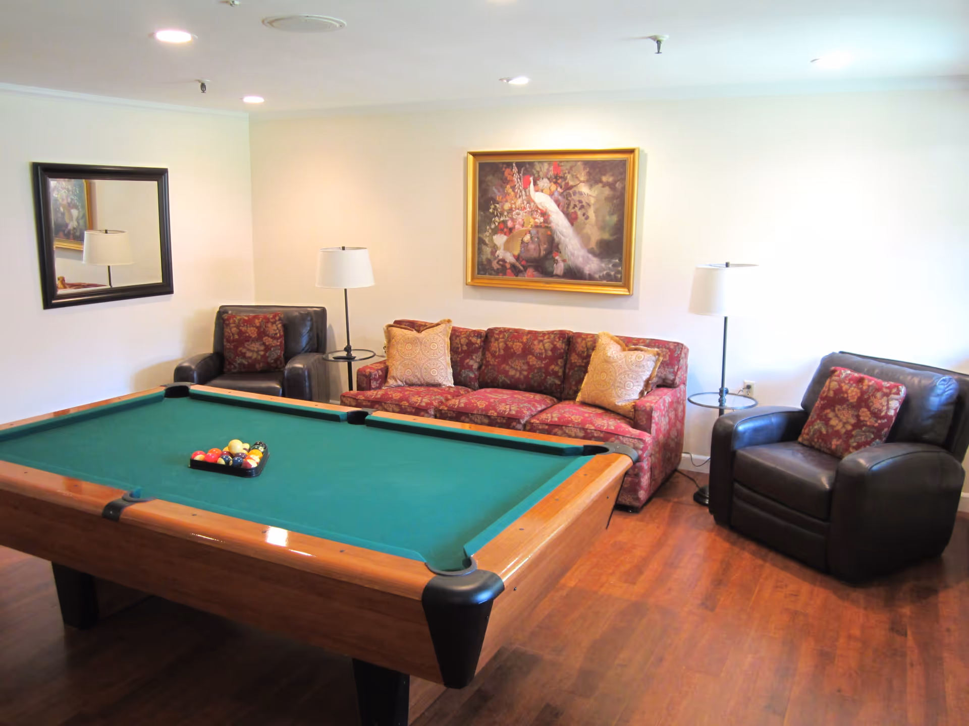 A cozy room with a pool table in the center, surrounded by a floral-patterned sofa and two dark leather armchairs with matching cushions. The room has wooden flooring, two floor lamps, a large mirror on one wall, and a framed painting of a white peacock above the sofa.