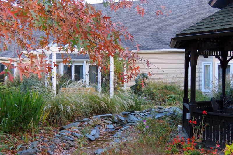 A peaceful outdoor garden area with a small rocky stream running through it, surrounded by various plants and flowers. There is a wooden gazebo on the right side with chairs inside, and a beige building with white columns and windows in the background. Autumn leaves hang from a tree branch in the upper left corner.
