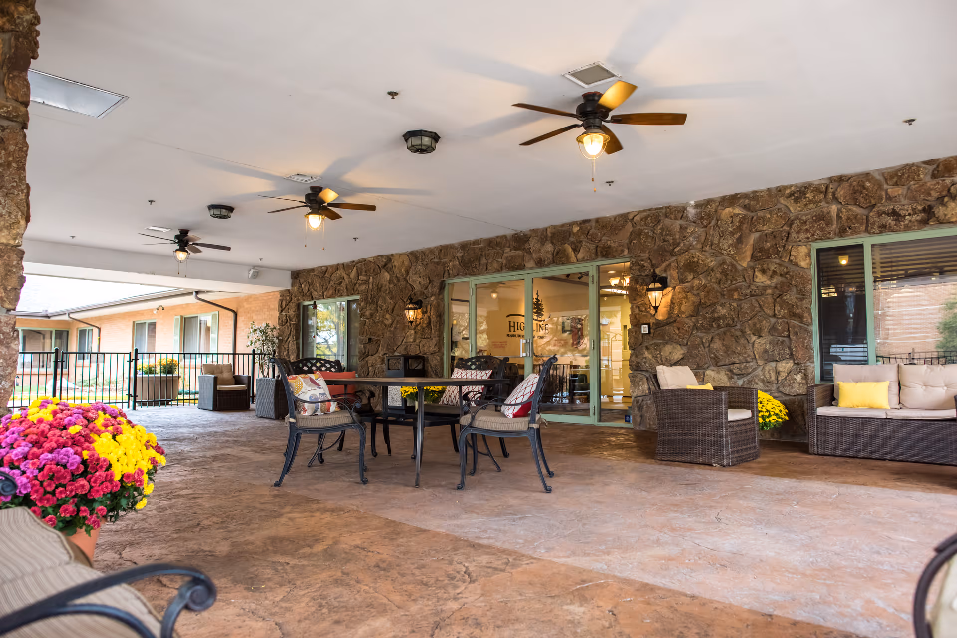 Covered outdoor patio area with stone walls, ceiling fans, and seating including chairs and sofas with cushions. There are colorful potted flowers and glass doors leading inside the building with the name Highline Post Acute visible through the glass.