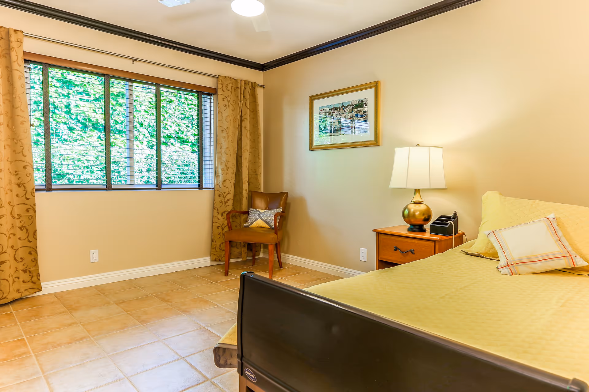 A bright bedroom featuring a bed with yellow bedding, a wooden nightstand with a lamp, a chair, and a large window with curtains.
