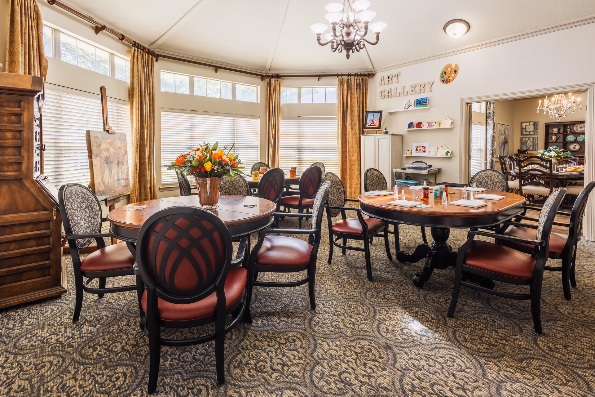A bright and inviting room with multiple round wooden tables surrounded by chairs with red cushioned seats and patterned backs. The room has large windows with blinds and gold curtains, a chandelier hanging from the ceiling, and a sign on the wall that reads 'ART GALLERY' with small art pieces displayed on shelves. There is a vase with colorful flowers on one of the tables and an easel with a painting near the window.