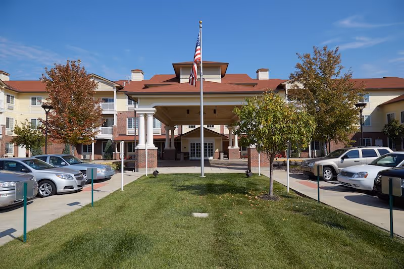 Front exterior view of Aventura Senior Living at Overland Park showing a three-story building with a covered entrance supported by white columns. There is a flagpole with an American flag in the center of a grassy area, flanked by small trees and parked cars on both sides under a clear blue sky.