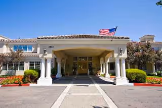 Front exterior view of Aegis Living Fremont facility with a covered entrance supported by white columns, an American flag flying on the roof, and landscaped bushes and trees around the building.
