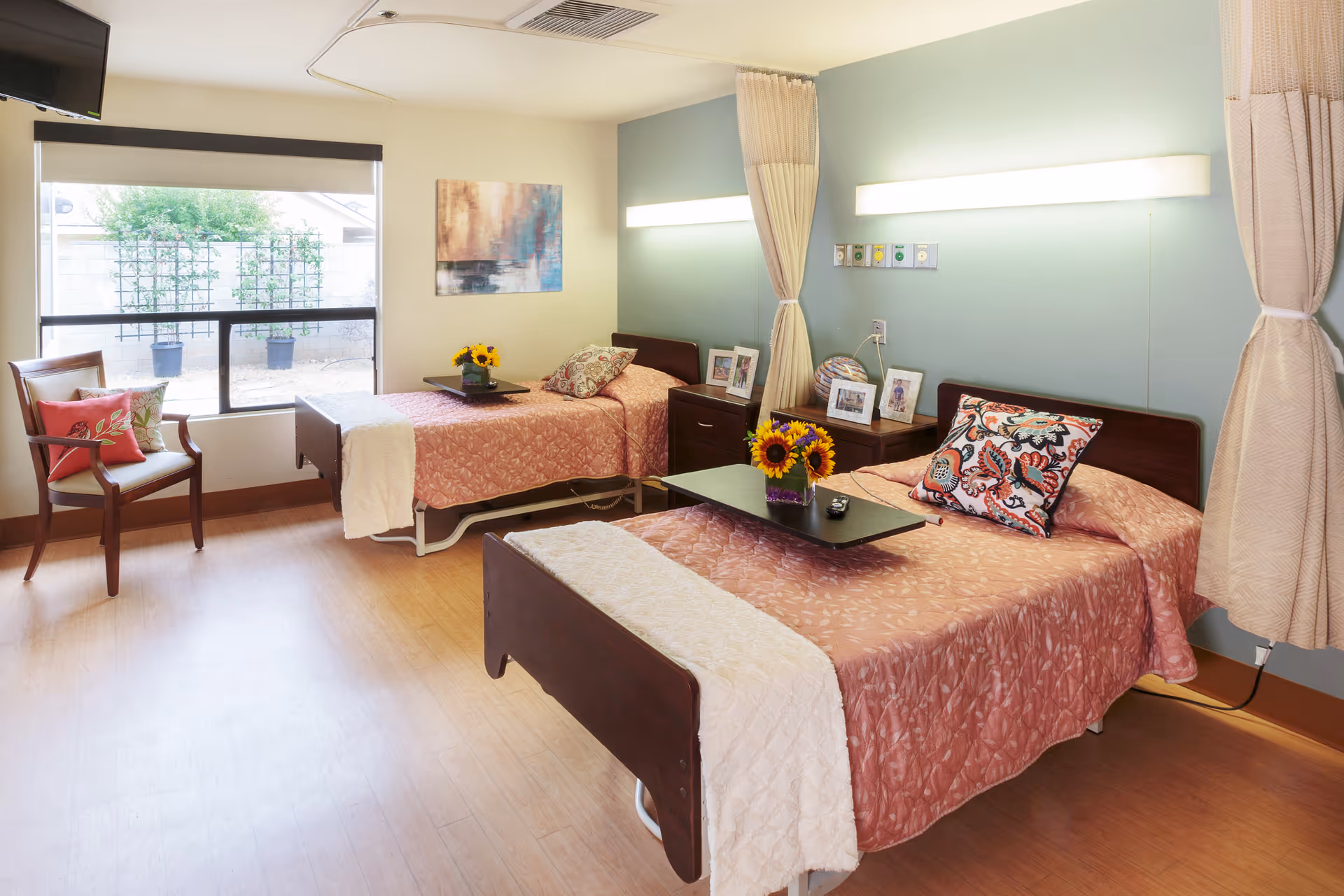 A bright and clean bedroom in a senior living facility with two single beds covered in pink bedspreads, each with a decorative pillow and a small black tray holding a vase of sunflowers. The room has light wood flooring, a large window with a view of an outdoor area, a wooden chair with cushions, and a wall-mounted TV. There are framed photos on the nightstands between the beds and soft lighting above each bed.