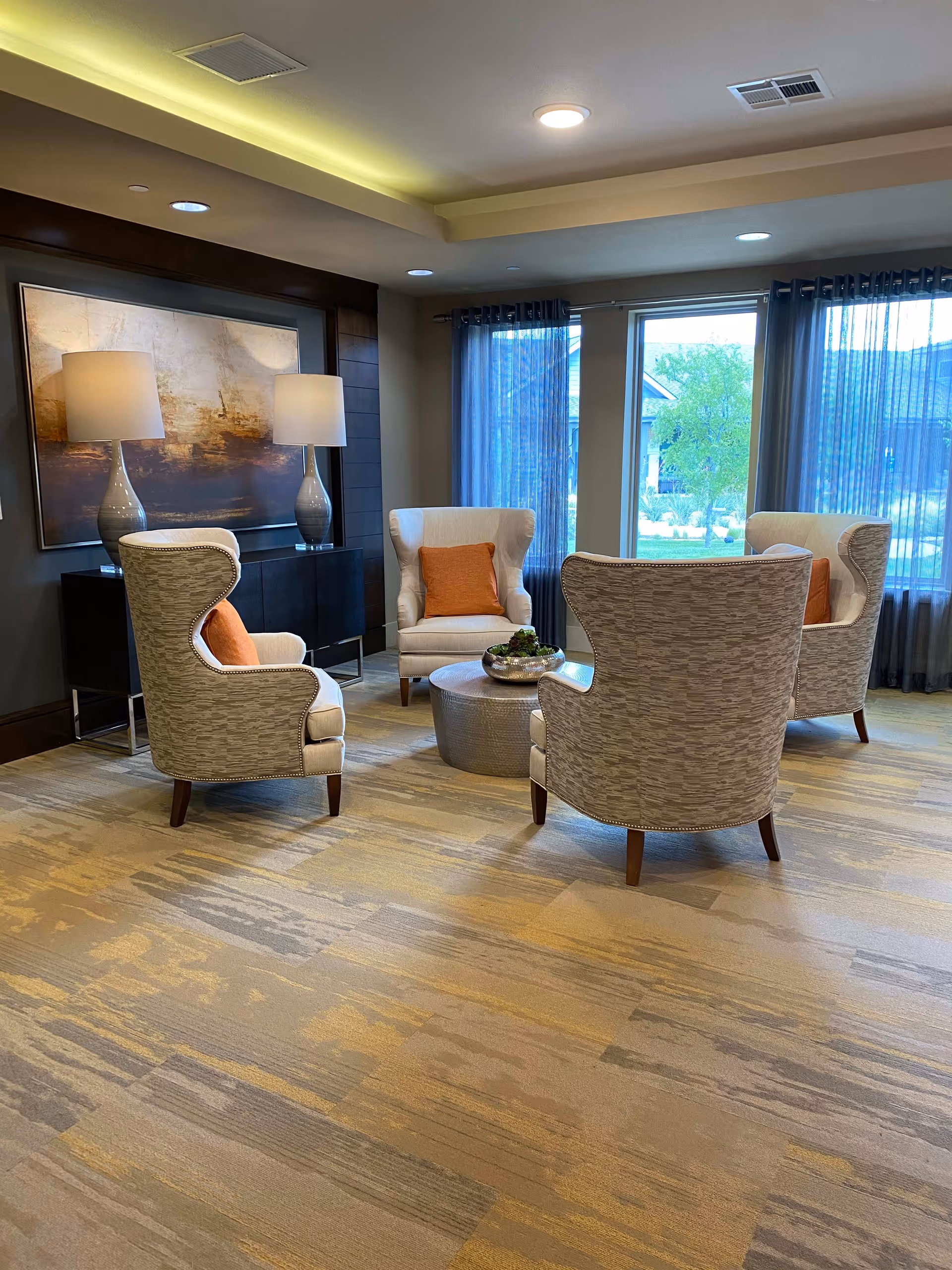 A cozy sitting area with four upholstered armchairs arranged around a round metallic coffee table. Each chair has an orange cushion. Behind the chairs is a dark wooden console table with two matching table lamps and a large abstract painting on the wall. Large windows with sheer curtains allow natural light to brighten the room.