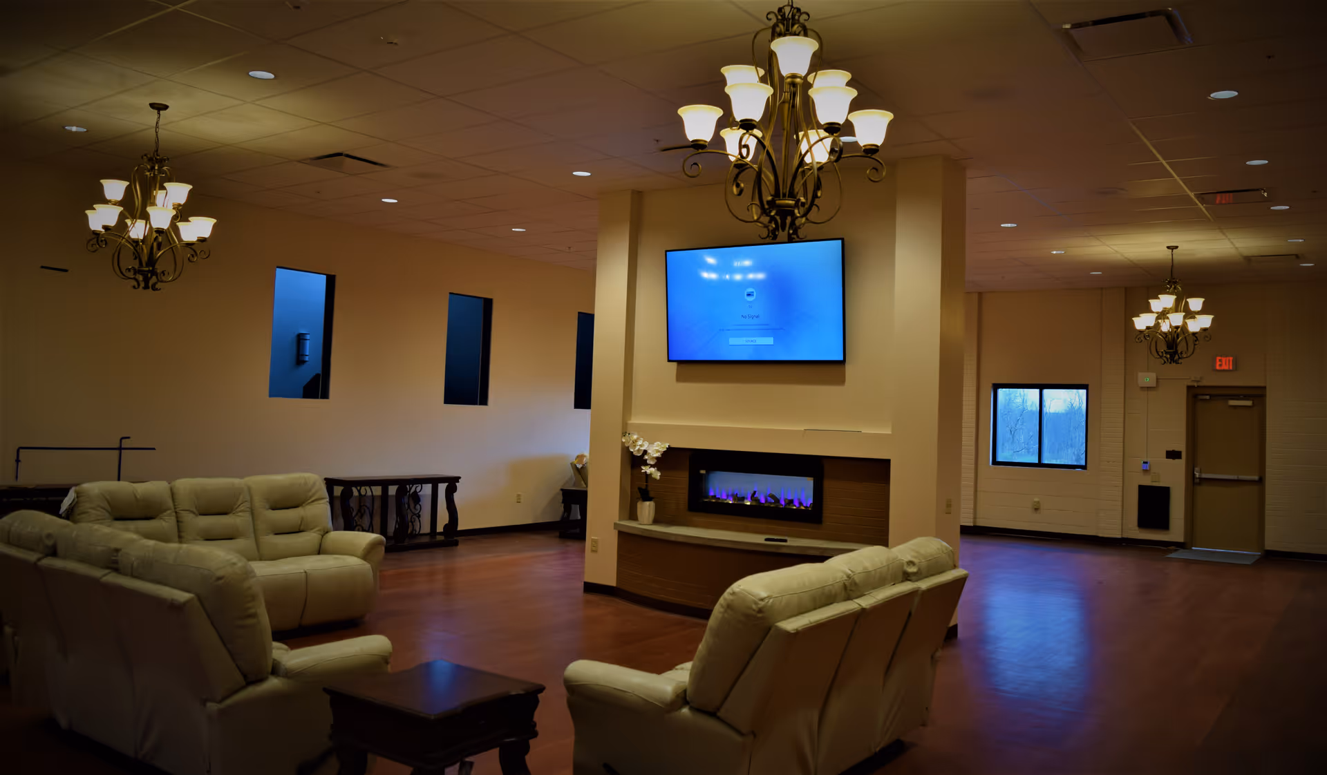 A spacious living room area with beige leather sofas arranged around a small wooden table. The room features a modern electric fireplace built into a central column, above which a flat-screen TV is mounted. The walls are light-colored with several small rectangular windows. Elegant chandeliers hang from the ceiling, and the floor is covered with wood-like flooring. There is a door and additional windows visible in the background.