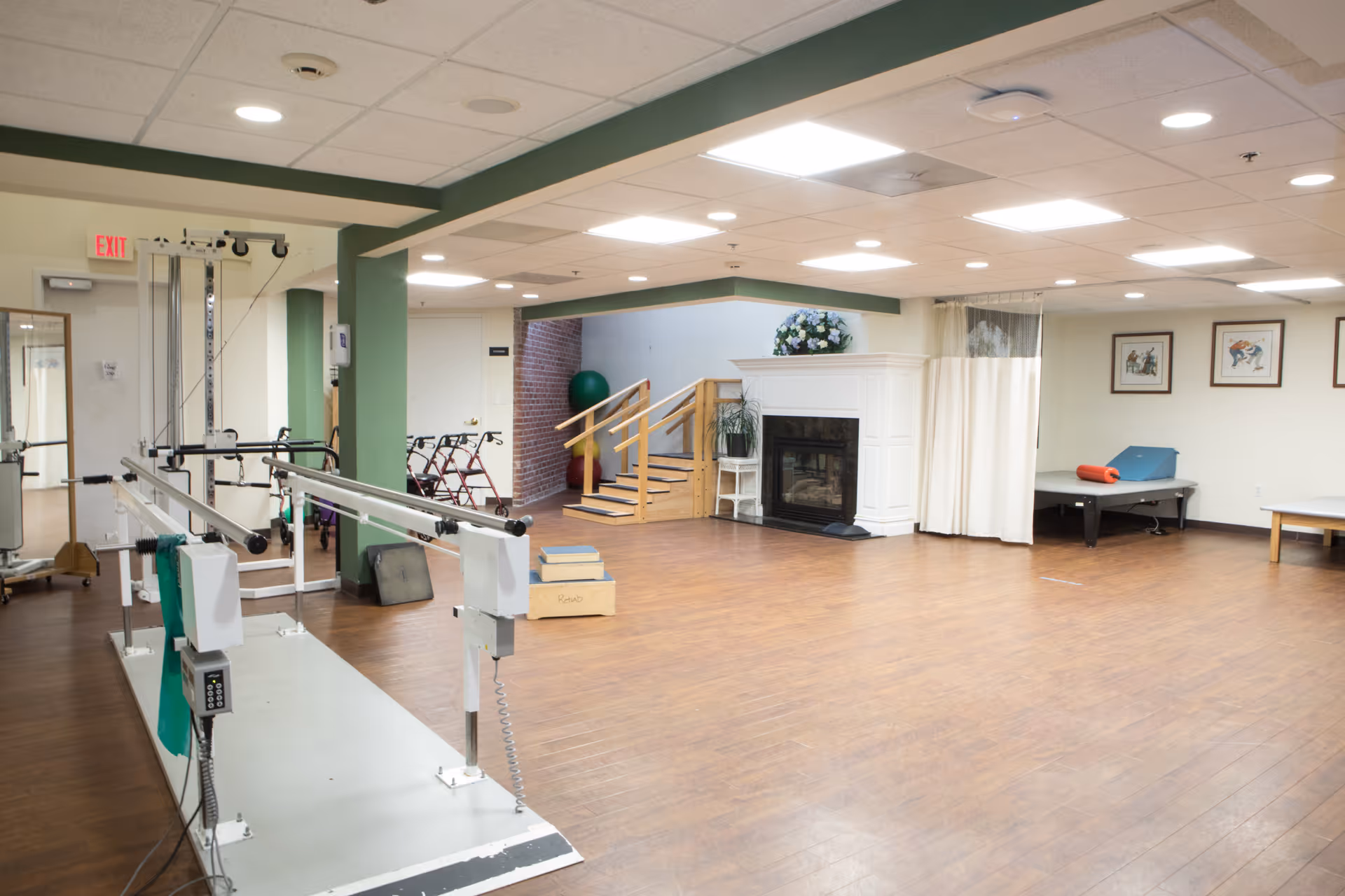 A spacious rehabilitation or physical therapy room with parallel bars for walking exercises, a small set of wooden stairs with handrails, a fireplace with a floral arrangement on top, exercise mats, and framed artwork on the walls. The room has wood flooring and bright overhead lighting.