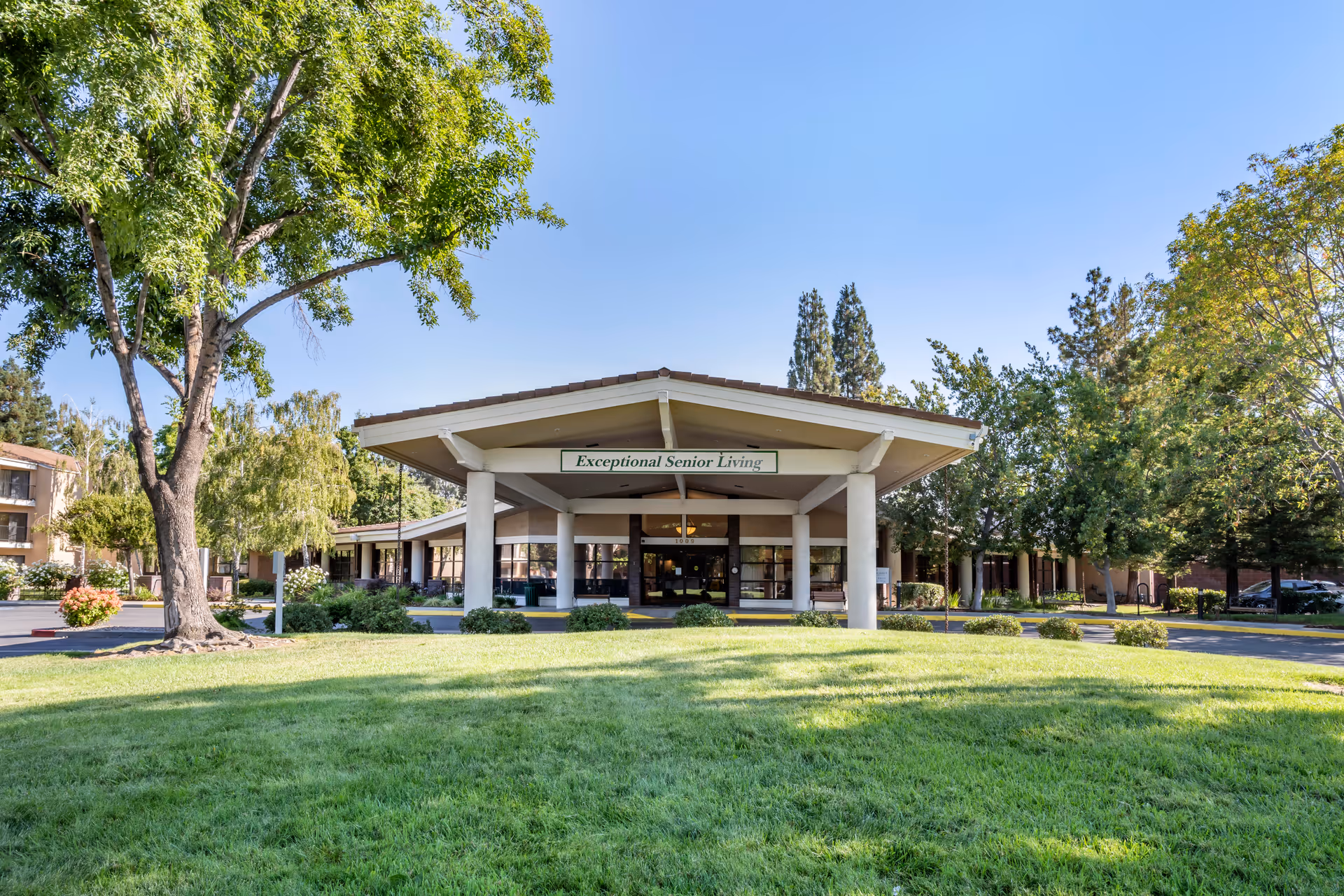 Front exterior view of Brookdale San Jose senior living facility with a covered entrance, surrounded by green grass, trees, and clear blue sky.