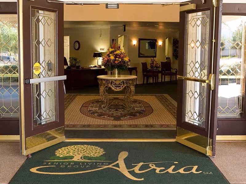 Open double glass doors with decorative gold trim leading into a warmly lit interior space featuring a round table with a large floral arrangement on a patterned rug. The floor mat outside the doors reads 'Senior Living Group Atria'.