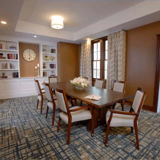 A well-lit dining room with a long wooden table surrounded by eight cushioned chairs. The table has a floral centerpiece and an open book. The room features patterned carpet, large windows with curtains, built-in white shelves with books and decorative items, and a wall clock.