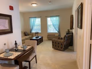 A cozy living room area with a beige carpet, a patterned armchair, a beige sofa, a wooden coffee table, and a small dining table set for two with placemats, plates, and a bottle of wine. Two windows with sheer blue curtains allow natural light into the room. The walls are decorated with framed artwork.