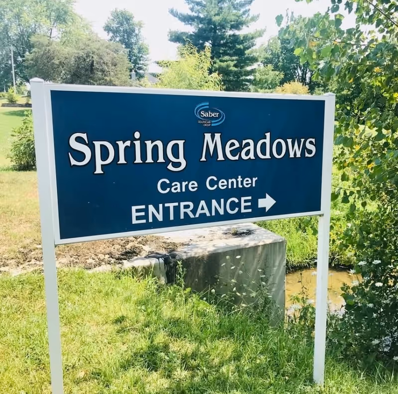 Outdoor sign for Spring Meadows Care Center with an arrow pointing to the entrance, surrounded by grass, trees, and bushes on a sunny day.