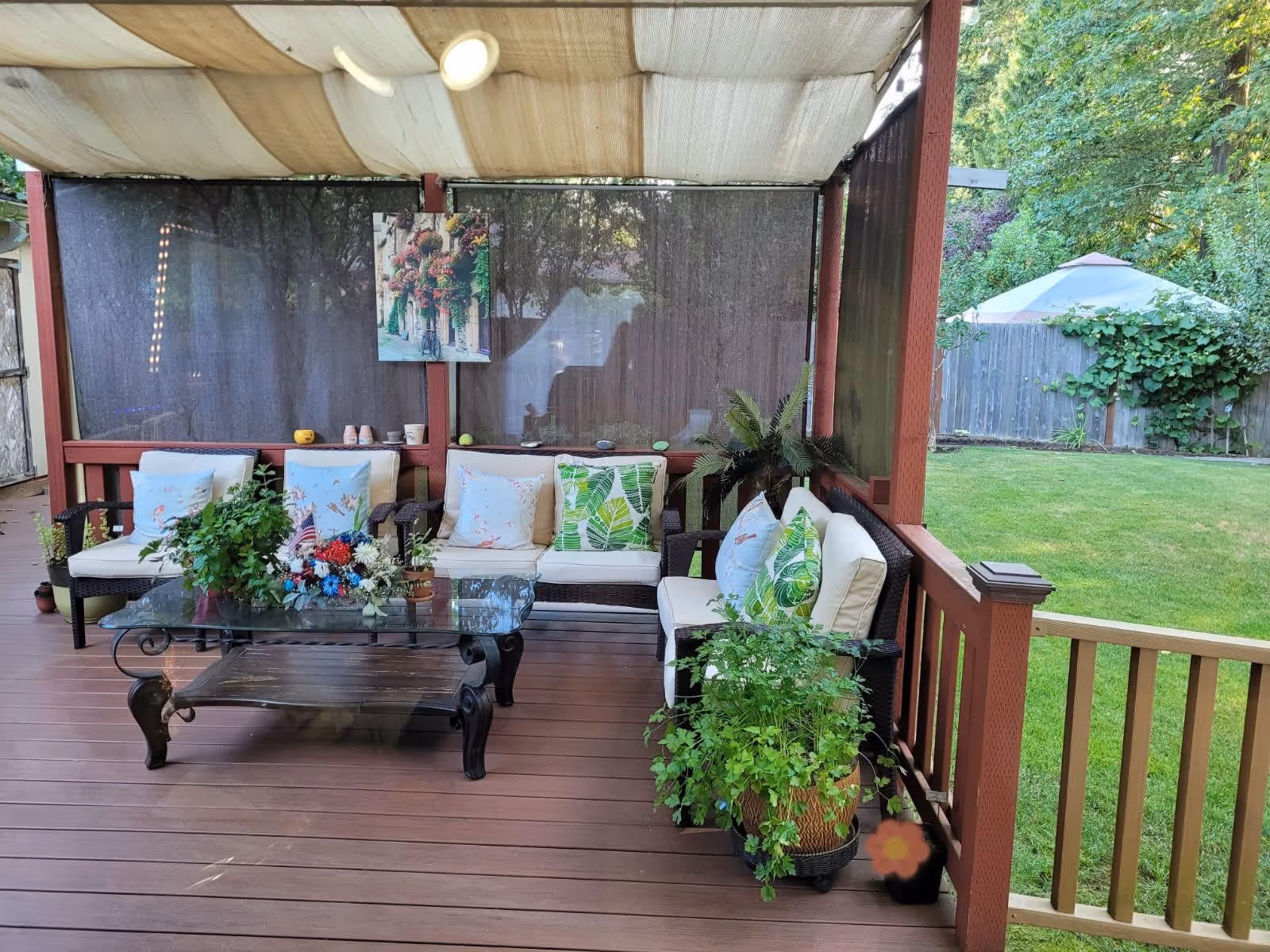 Covered outdoor patio area with cushioned wicker seating, a glass coffee table adorned with flower arrangements and potted plants, surrounded by a wooden deck and overlooking a green lawn with trees and a gazebo in the background.