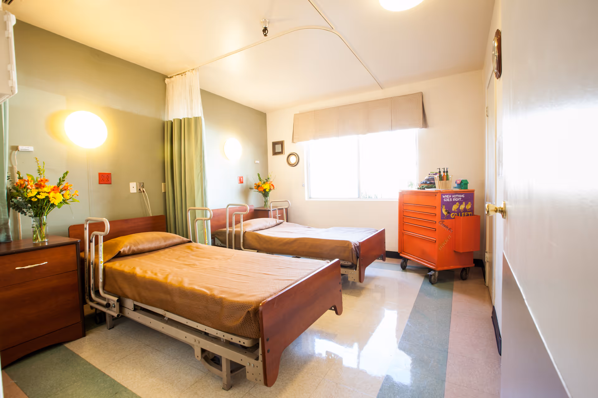 A bright nursing center room with two hospital beds covered in brown bedding. Each bed has metal side rails and is positioned against a green accent wall with round wall lights above. There are two wooden nightstands with flower vases on each side of the beds. A large window with a beige valance lets in natural light. An orange rolling cart with various items and a humorous sign is near the window. The floor has a patterned linoleum design.