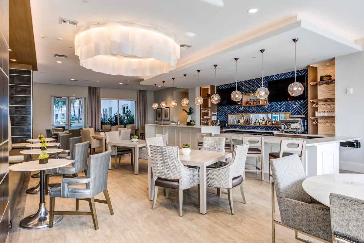 Bright modern dining and bar area with multiple tables, upholstered chairs, pendant lights, and a service counter.
