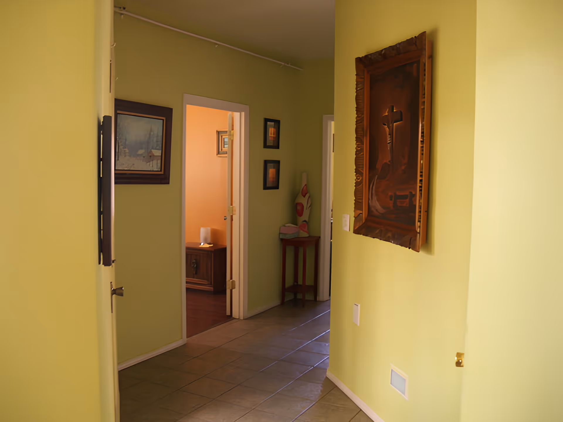 A light-green interior hallway with a tiled floor and framed artwork leading to a doorway into another room.