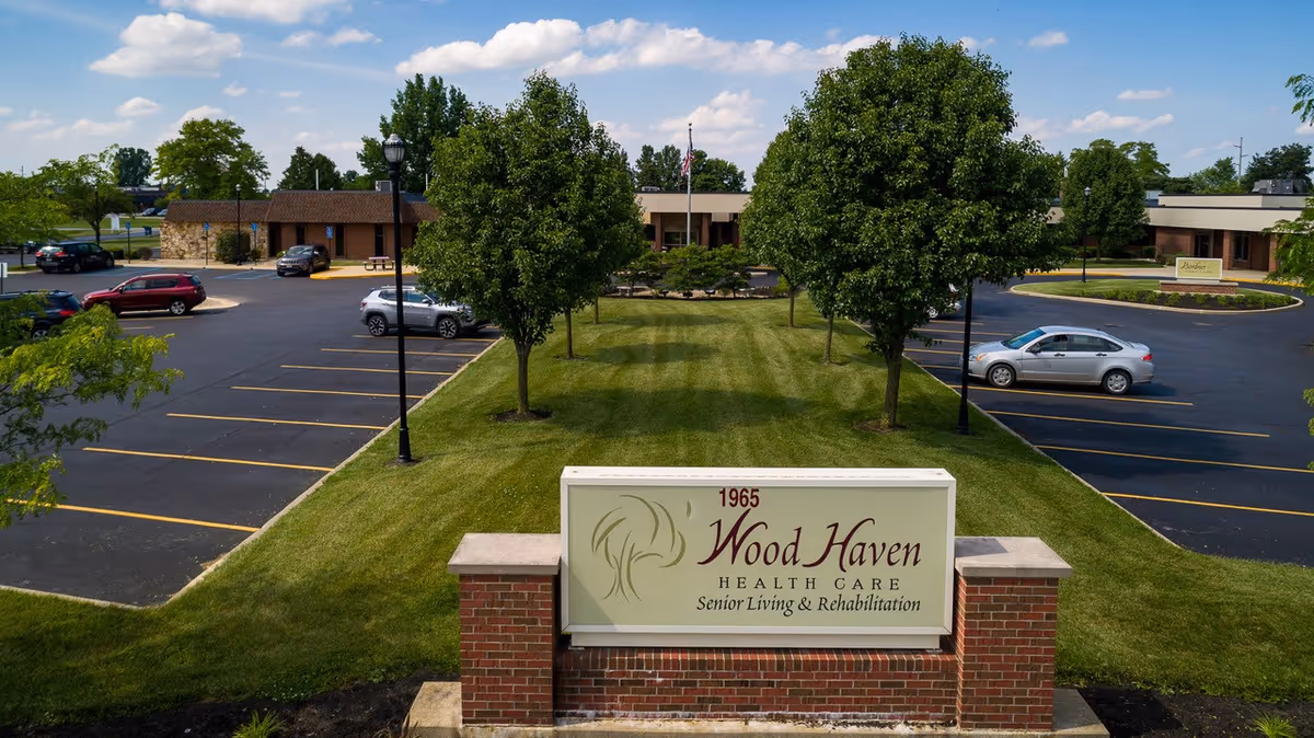 Exterior view of Wood Haven Health Care facility showing a parking lot with several cars, a green lawn with trees, and a sign in the foreground that reads '1965 Wood Haven Health Care Senior Living & Rehabilitation'. The building is single-story with a brown roof and surrounded by trees under a partly cloudy sky.
