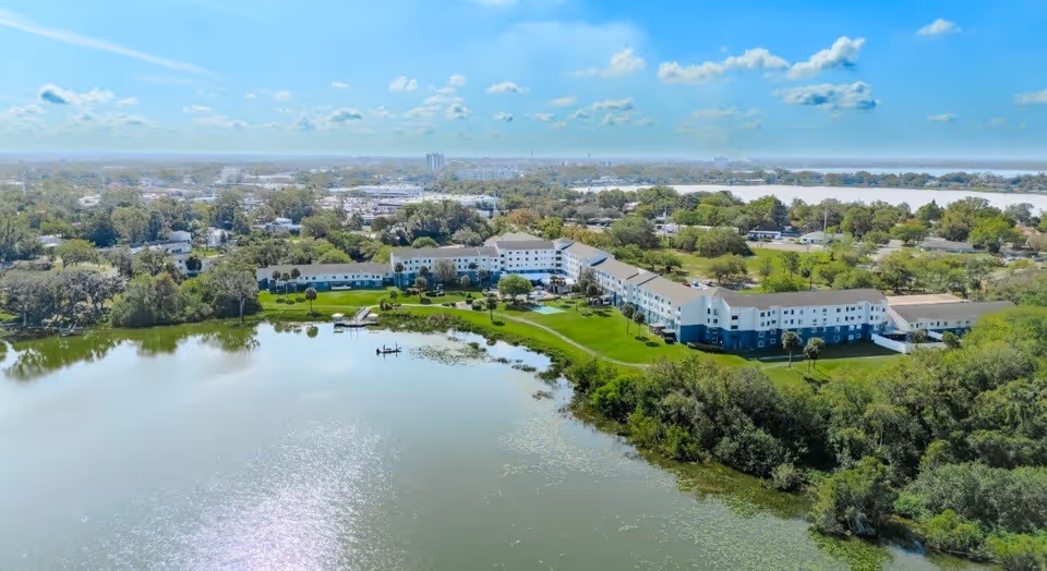Aerial view of a large senior living facility named Spring Haven situated next to a lake with green lawns and trees surrounding the building. The sky is clear with some clouds, and the area around the facility includes residential and commercial buildings.