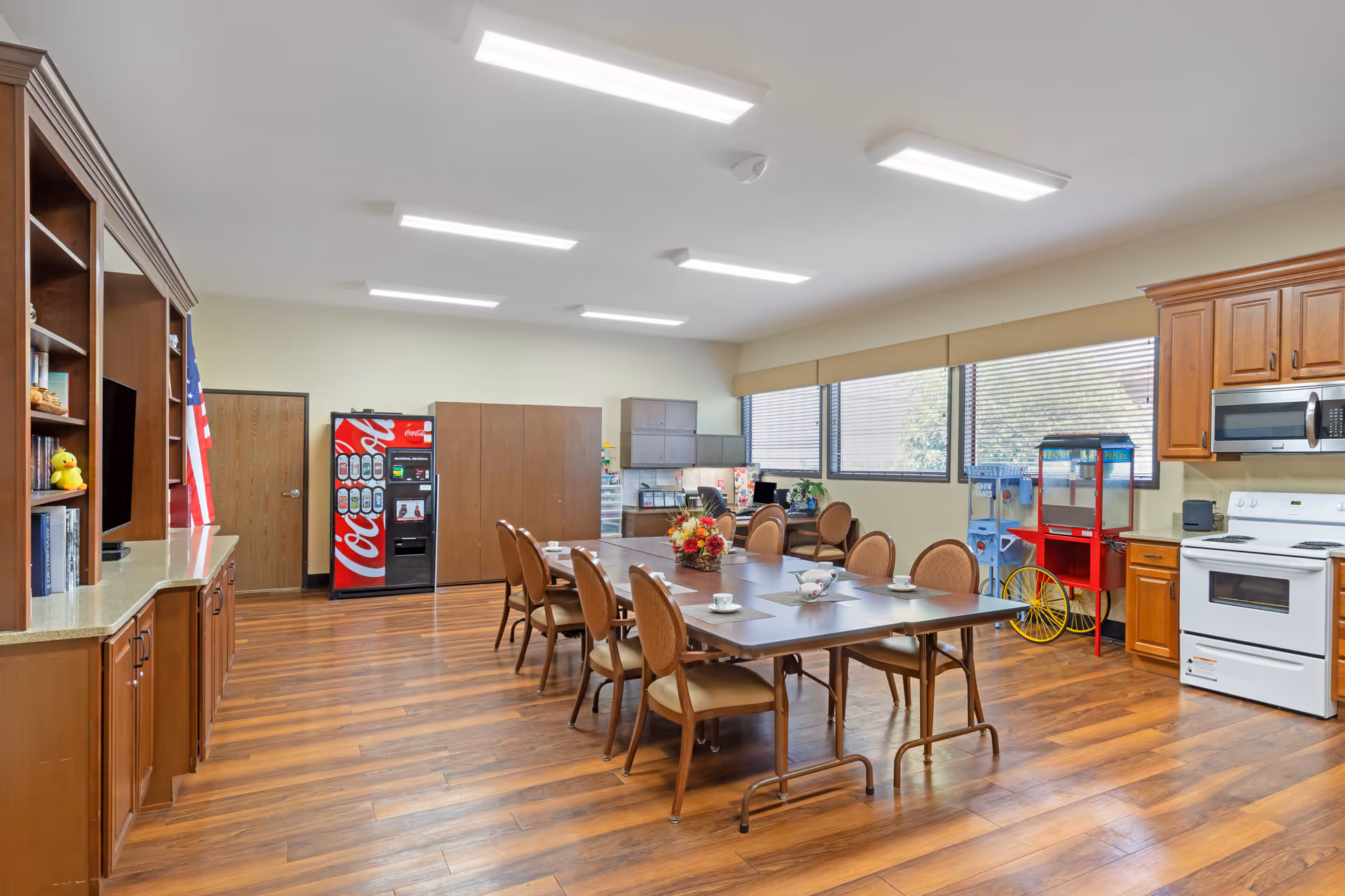 A spacious room with wooden flooring and cabinetry, featuring a long table with eight chairs arranged around it. The table is set with cups and saucers, and a floral centerpiece. The room includes a kitchen area with a white stove, microwave, and wooden cabinets. There is a Coca-Cola vending machine, a popcorn machine, and a water dispenser near the windows, which have blinds partially open to let in natural light.