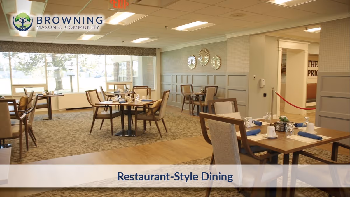 A bright and spacious restaurant-style dining room with several wooden tables and cushioned chairs arranged neatly. Each table is set with white cups, glasses, napkins, and silverware. Large windows allow natural light to fill the room, and decorative wall accents are visible. The room has a warm and inviting atmosphere.