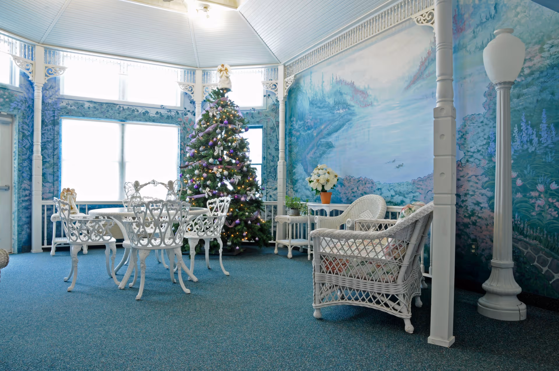 A bright indoor sitting area decorated for Christmas with a Christmas tree adorned with lights and ornaments. The room features white wicker furniture including chairs and a table, with floral cushions. The walls are painted with a scenic mural of a lake and trees, and there is a tall white lamp post inside the room. Large windows allow natural light to fill the space.