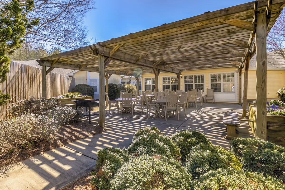 Outdoor patio area at TerraBella Newton with a wooden pergola providing partial shade over several tables and chairs. The patio is surrounded by bushes and plants, with a wooden fence on one side and a building with multiple windows in the background.